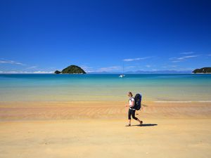 woman wearing backpack walking along a golden beach with blue sea and sky and an offshore island