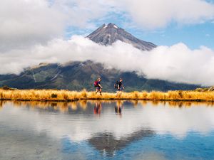 two people walking beside a lake with a volcano behind that's surrounded by white clouds