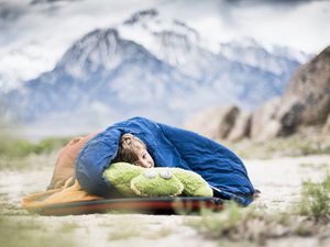 boy snuggled up in sleeping back outside with mountain in background