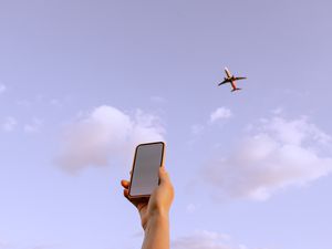 hand holding a smartphone against a purple-ish sky with an airplane flying above