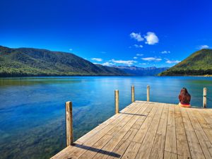 blue lake and sky with forested hills and woman sitting on a wooden jetty