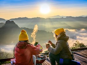 two people in yellow beanies enjoying a hot drink while looking out at mountains and fog below