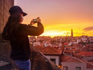 Teenage brunette girl dressed in black and wearing a cap wearing a surgical mask during a pandemic, sightseeing, taking photos of the beautiful sunset over the houses in Porto