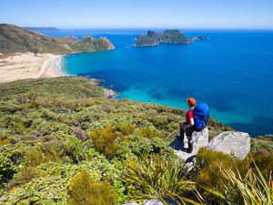 hiker wearing red hat and blue backpack looks out over the sea from a high rocky point