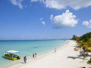 People walking on white sand beach near clear blue water with covered green and yellow boat in the foreground at Negril, Jamaica.