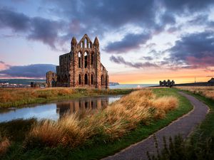 ruins of Whitby abbey at sunset on a cloudy day. The abbey is an field with over grown grasses, a small pond, and a narrow paved footpath