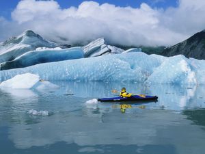 A kayaker paddles on serene water in front of a ice-blue glacier.