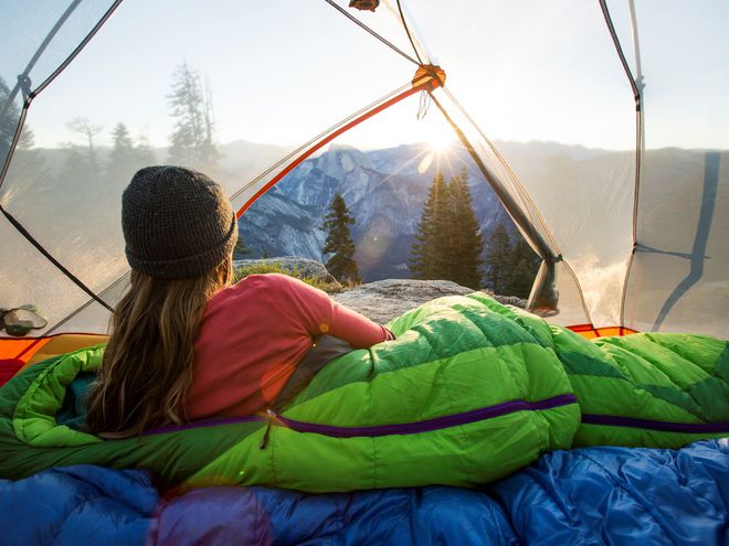 woman wearing a beanie hat lying in a tent in a green sleeping bag with mountain views outside