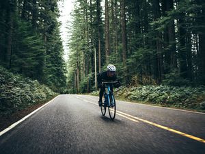 A man cycling in a beautiful Oregon state forest setting on the road to Larch Mountain. He has a bright blue street bike that stands out from the green of the surrounding trees. He looks up the road ahead as he pedals hard downhill, getting in some training for upcoming races. Motion blur on the road as he speeds down.