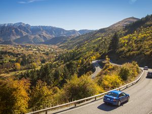 blue car drives down winding road with trees and mountains in the background