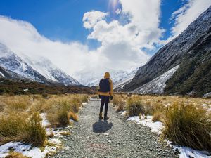 person wearing yellow jacket and black backpack walks along a stony path with mountains and clouds around
