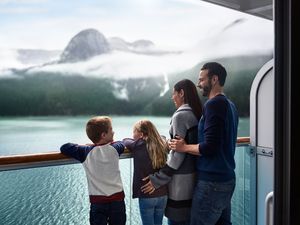Brunette man and woman standing on a cruise ship balcone with their young chidlren (a blonde boy and girl). The family is looking at mountains and rippling water