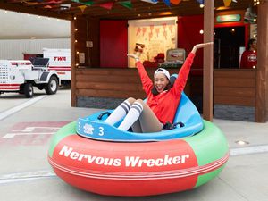 Smiling lightskinned black woman sitting into a bumper car with her hands in a "V" shape. The bumper car is in front of a small outdoor stall