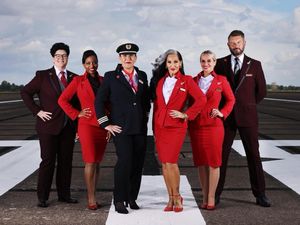 Group of six people (including Michelle Visage) posing on an airport tarmac in red and burgundy suits. The two people on either end are wearing burgundy pant suits. The person in the middle left is in a pilots uniform and heels, and the other three people are in red skirt suits with matching heels.
