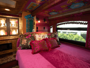 Full size bed in Dolly Parton's old tour bus. The bed has bright pink linens and many brocade pillows. There is a window to the right of the bed with pink fringe curtains. You can see bushes outside the window. There is a cabinet of four blonde wigs on the back wall of the tour bush. 